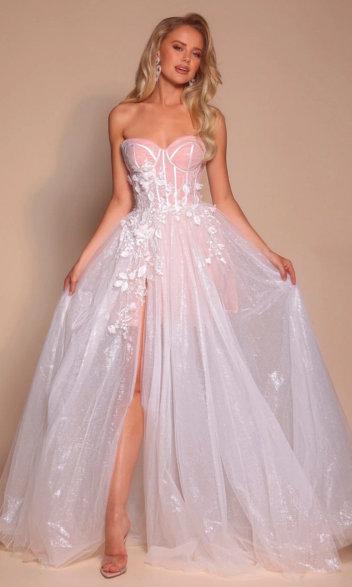A woman poses against a beige background in the Portia and Scarlett PS26078 long formal dress, featuring an ivory strapless sheer corset bodice with floral appliqué, a glitter tulle skirt, and a high slit.